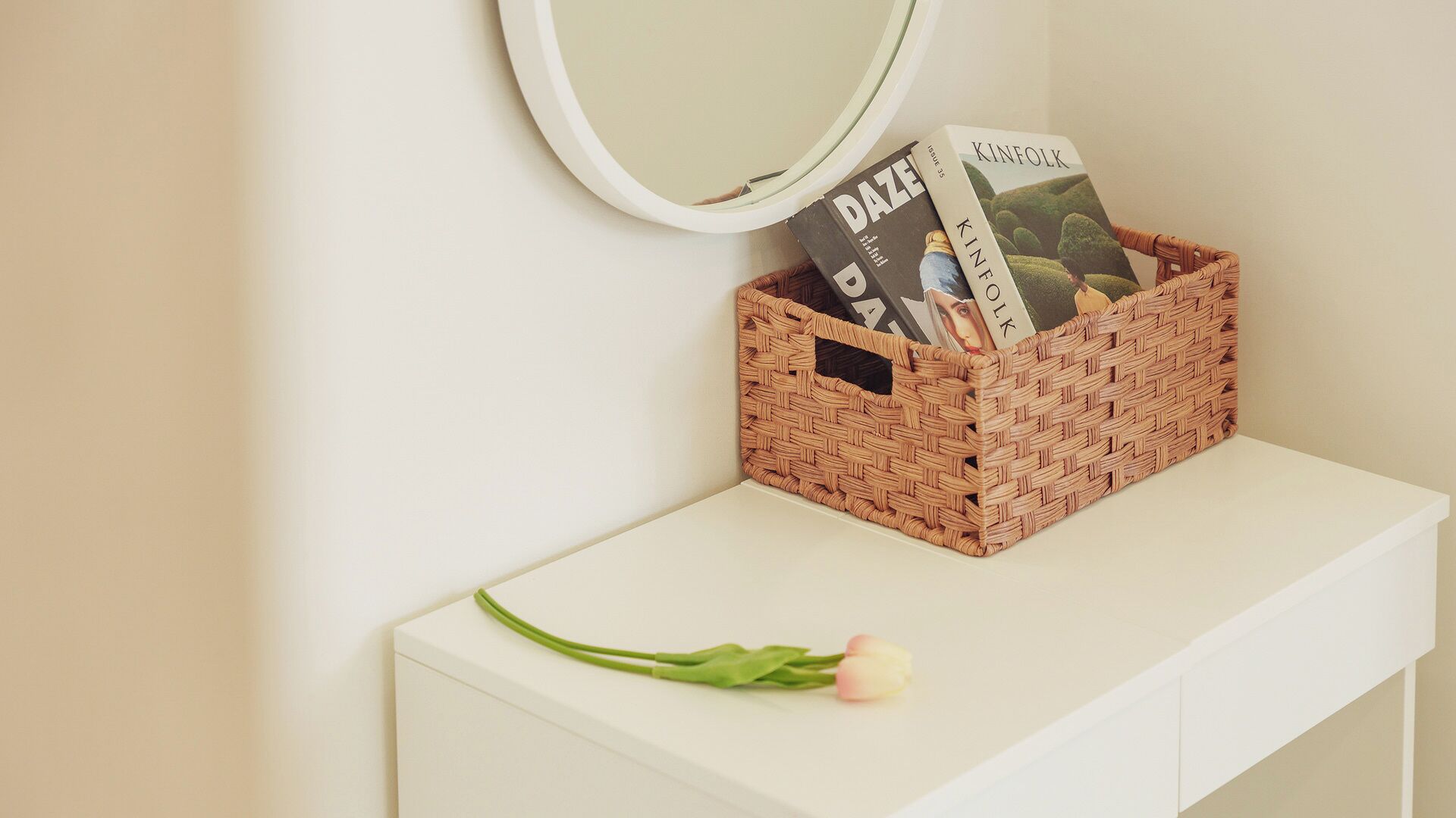 A white bedside table with a woven basket containing magazines and a single pink flower.