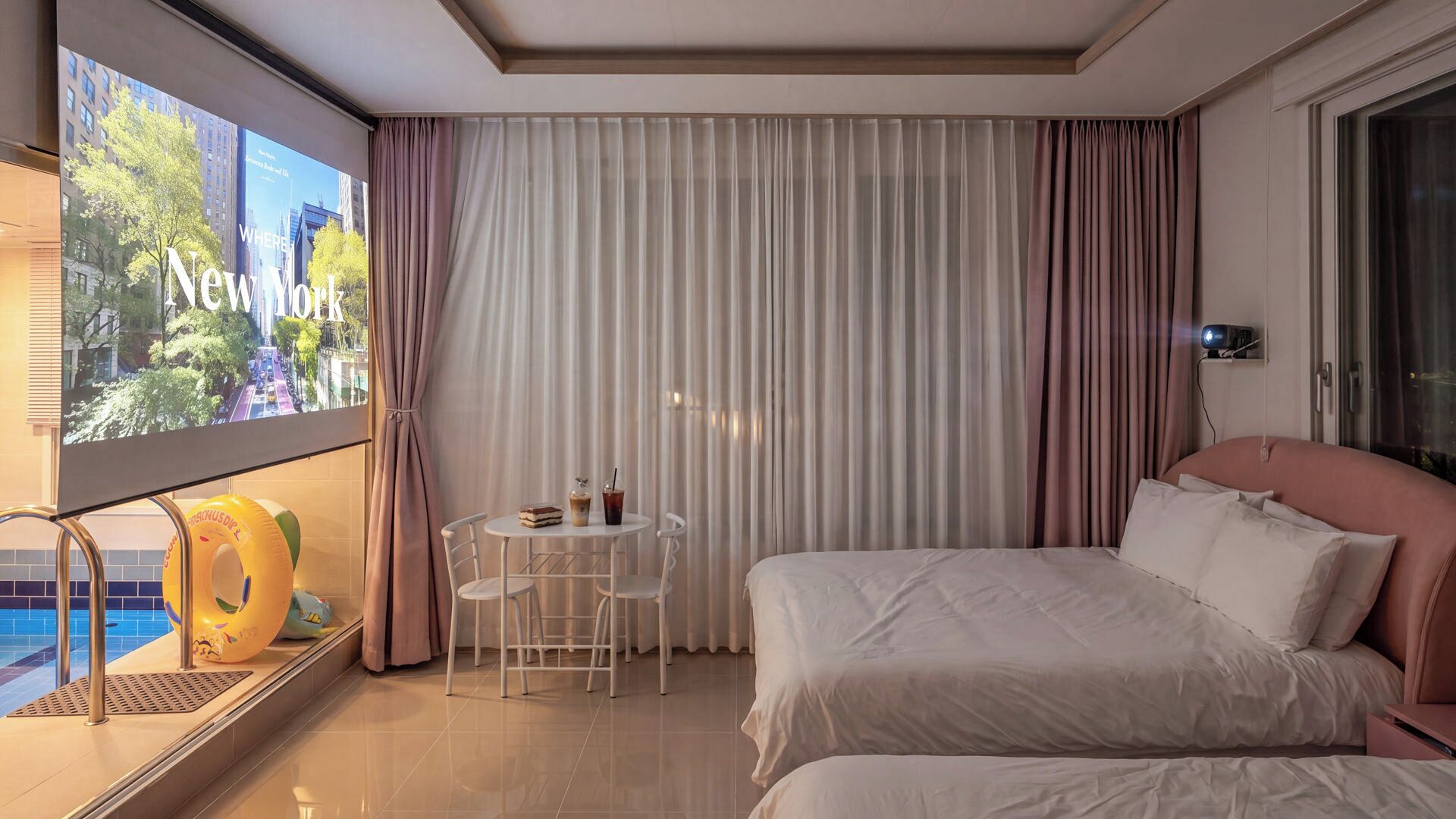 A hotel room with a bed, a small table with a drink, a chair, and a window with curtains.