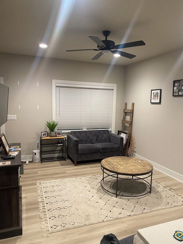 A living room with a dark grey sofa, a round coffee table, a ceiling fan, a window with blinds, and a small plant on a side table. - 3-bedroom townhome in awesome Ankeny with WiFi, fitness room, AC (Ankeny)