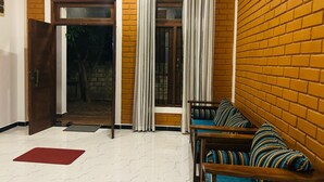 A room with a brick wall, a wooden door, a window with curtains, a sofa with patterned cushions, and a red mat on the floor. - Eco Brick Transit villa  AC in Katunayake (Katunayake)