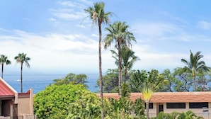 A coastal hotel with palm trees, a view of the ocean, and a red-roofed building. - Haleakala Shores B512: Luxe top-floor with dual master suites. Steps to beach. (Kihei)