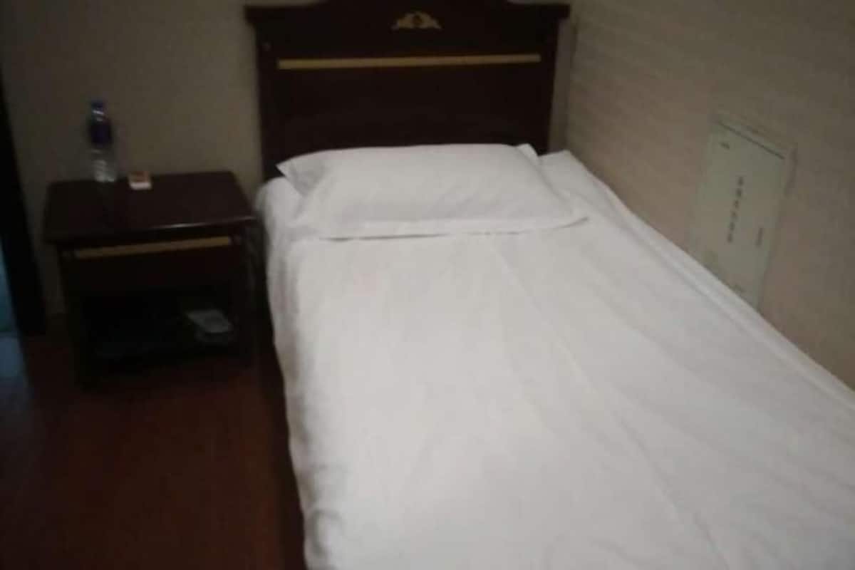 A single bed with white bedding, a wooden headboard, a small bedside table with a water bottle, and a wall-mounted electrical outlet.