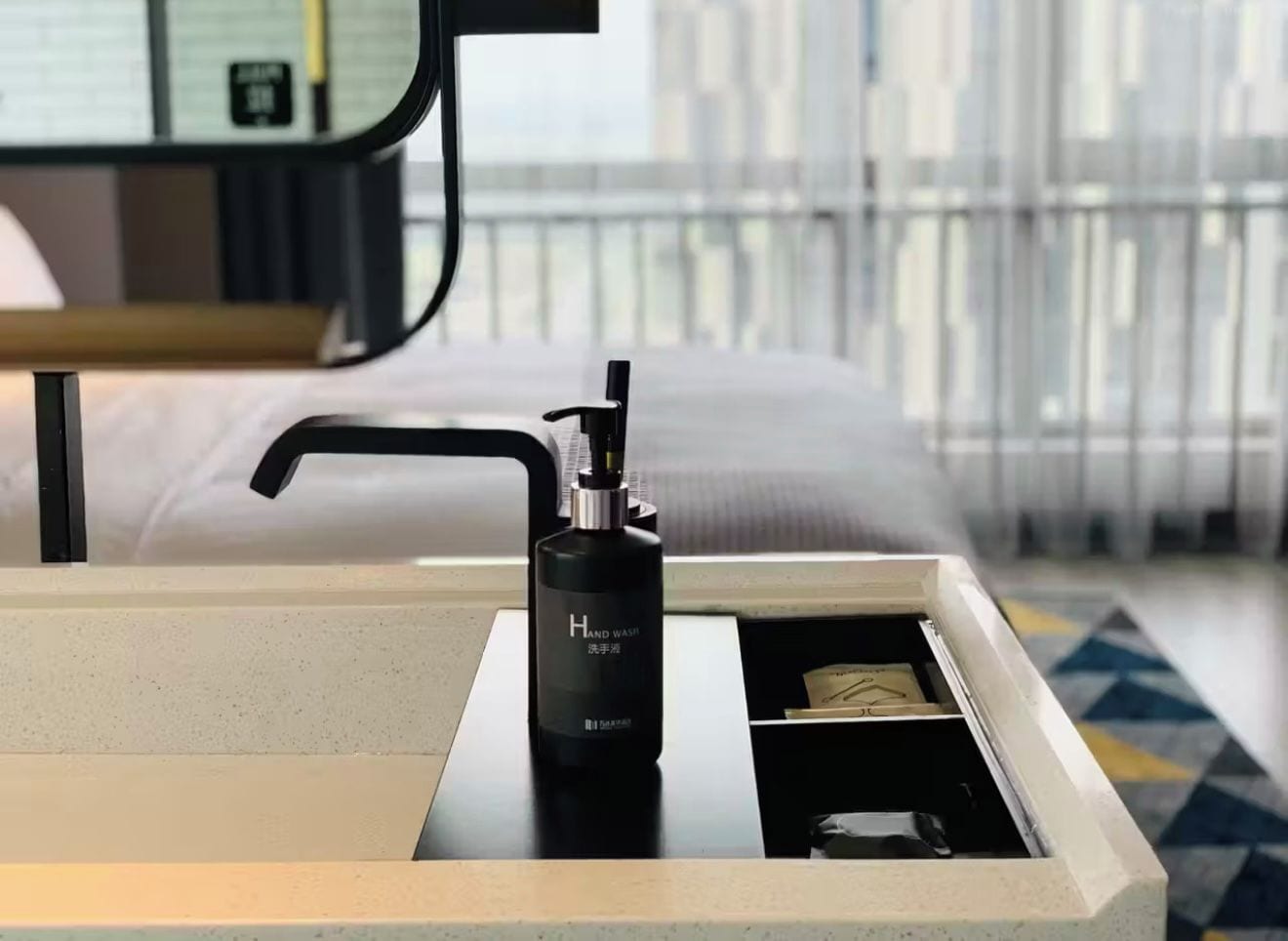 A hotel room with a bedside table, a black bottle of hand sanitizer, and a bed with white bedding.