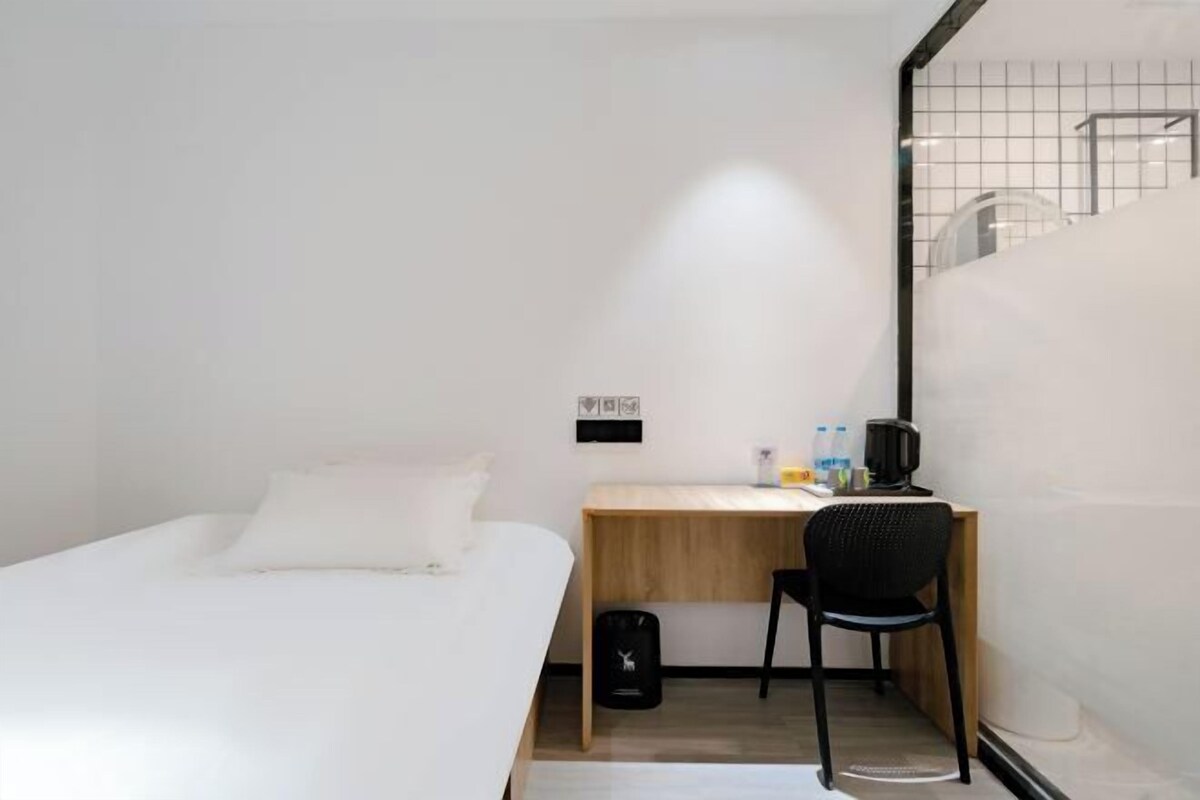 A small, modern hotel room with a single bed, a wooden desk, a chair, and a small trash bin.