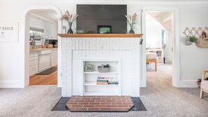 Ein Wohnzimmer mit Kamin aus weißem Backstein, einem Fernseher darüber und einem Bücherregal mit Büchern und Dekorationsobjekten.
