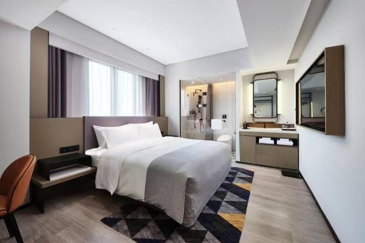A modern hotel room with a large bed, a desk, a chair, a bathroom with a shower, and a mirror.