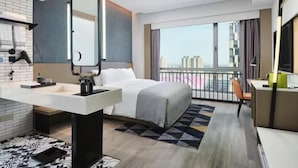 Una habitación de hotel moderna con una cama grande, un escritorio, una silla, un baño con espejo y vistas a la ciudad.