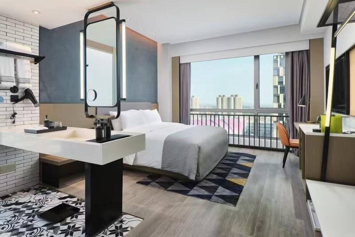 A modern hotel room with a large bed, a desk, a chair, a bathroom with a mirror, and a view of the city.
