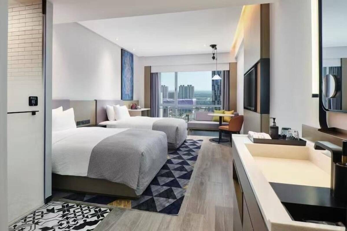A modern hotel room with two beds, a sofa, a desk, and a large window offering a city view.