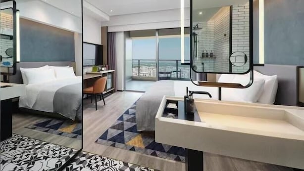 Una habitación de hotel moderna con una cama grande, un escritorio, un baño con un espejo amplio y un balcón con vista a la ciudad.
