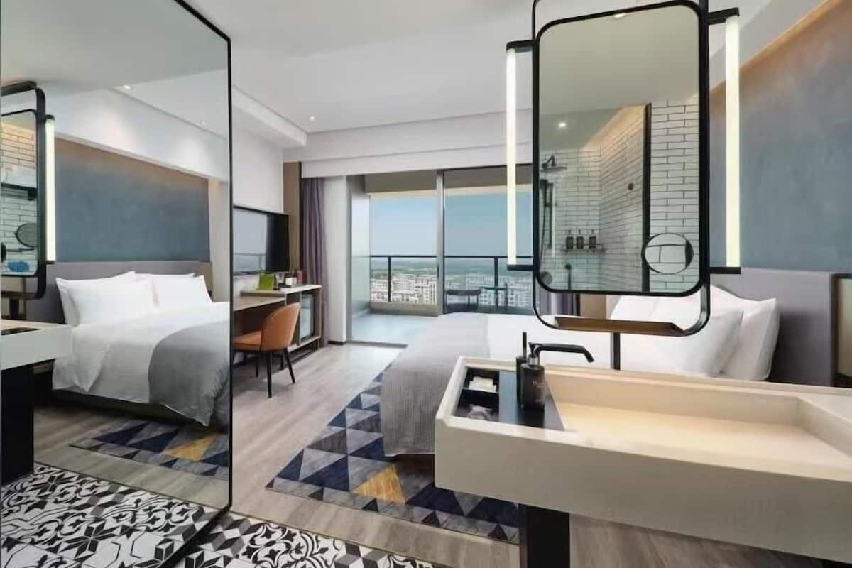 A modern hotel room with a large bed, a desk, a bathroom with a large mirror, and a balcony with a city view.