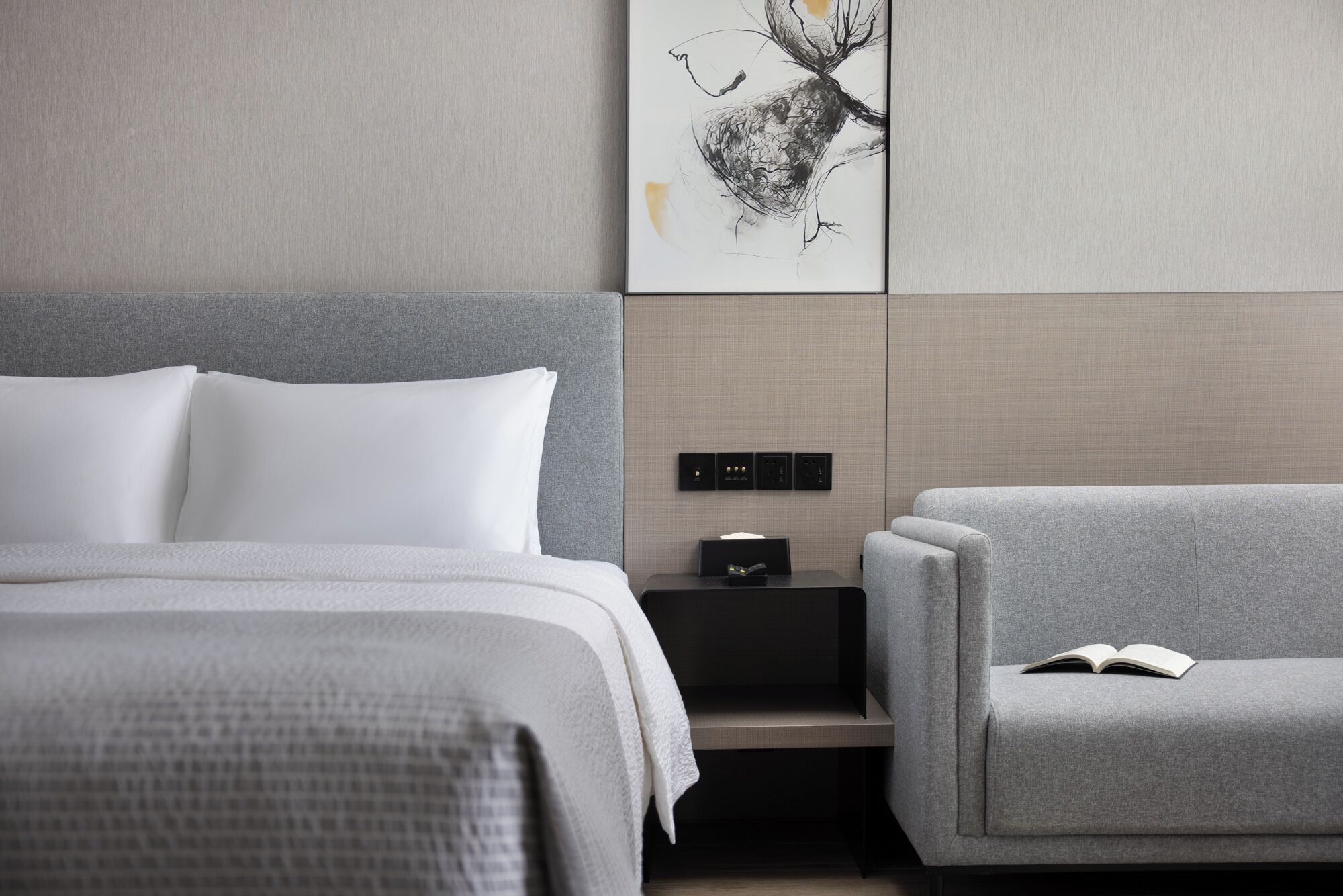 A modern hotel room with a bed, a gray sofa, a bedside table with a book, and a wall-mounted control panel.