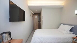 Een hotelkamer met een bed, een aan de muur bevestigde televisie, een nachtkastje met een waterkoker, en een deur naar een andere kamer.