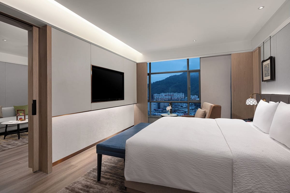 Een moderne hotelkamer met een groot bed, een flatscreen televisie aan de muur, uitzicht op een stadsgezicht door grote ramen en een zithoek met een tafel en stoelen.