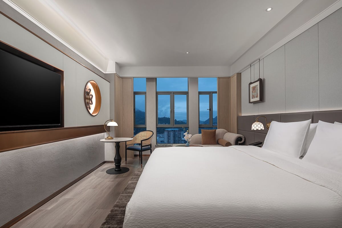 Een moderne hotelkamer met een groot bed, een flatscreen televisie, een klein rond tafeltje met een lamp, een stoel en uitzicht op de stad door grote ramen.