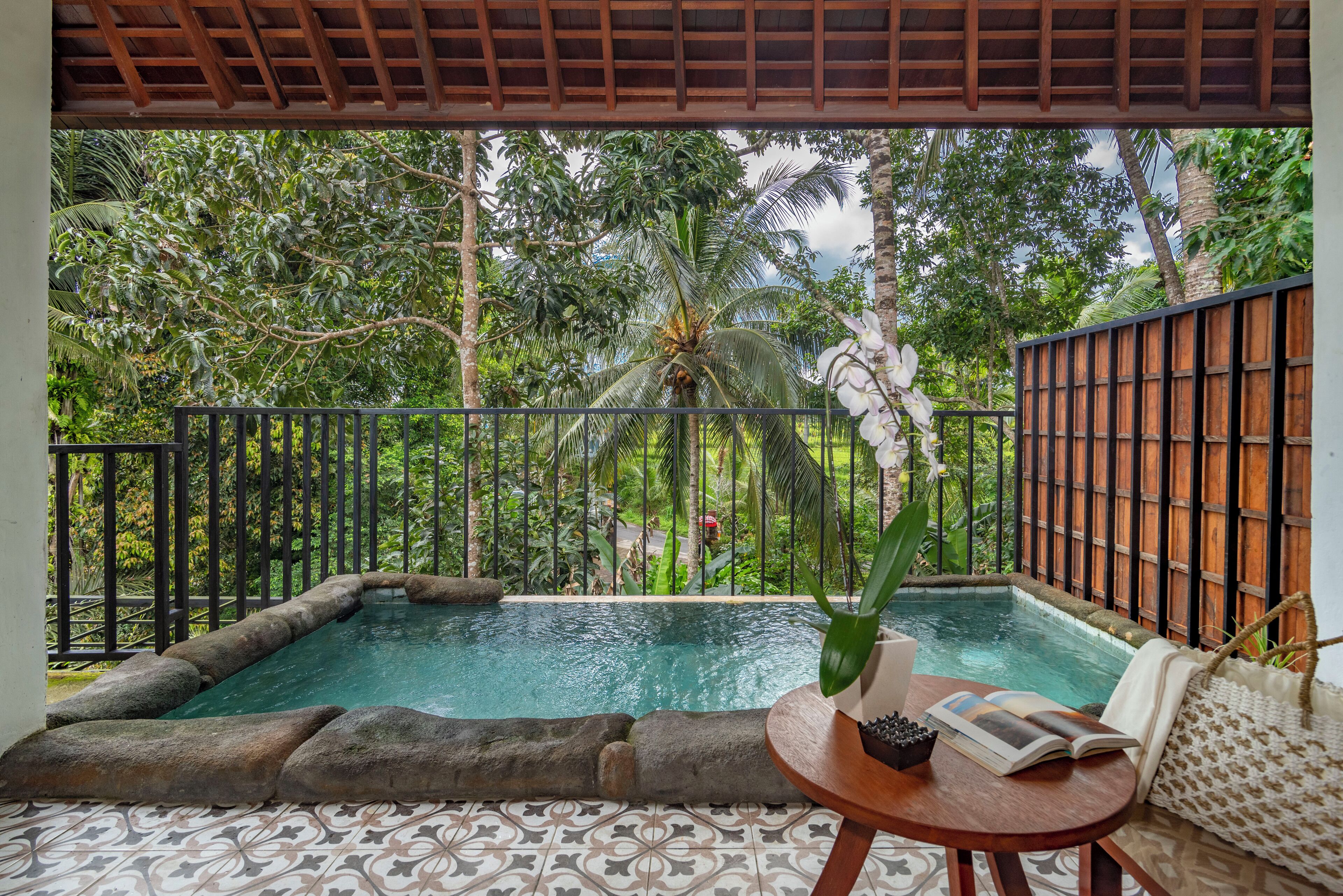 A pool area with a tiled floor, a wooden table with a book and a plant, and a view of lush greenery and a palm tree.