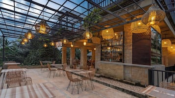 An outdoor patio area with tables and chairs, a bar area with shelves of bottles, and hanging lights.