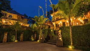 A well-lit courtyard with a staircase leading to a balcony, surrounded by palm trees and stone pillars. - Hoshi Jungle Resort by Dhananjaya Hospitality (Tegallalang)