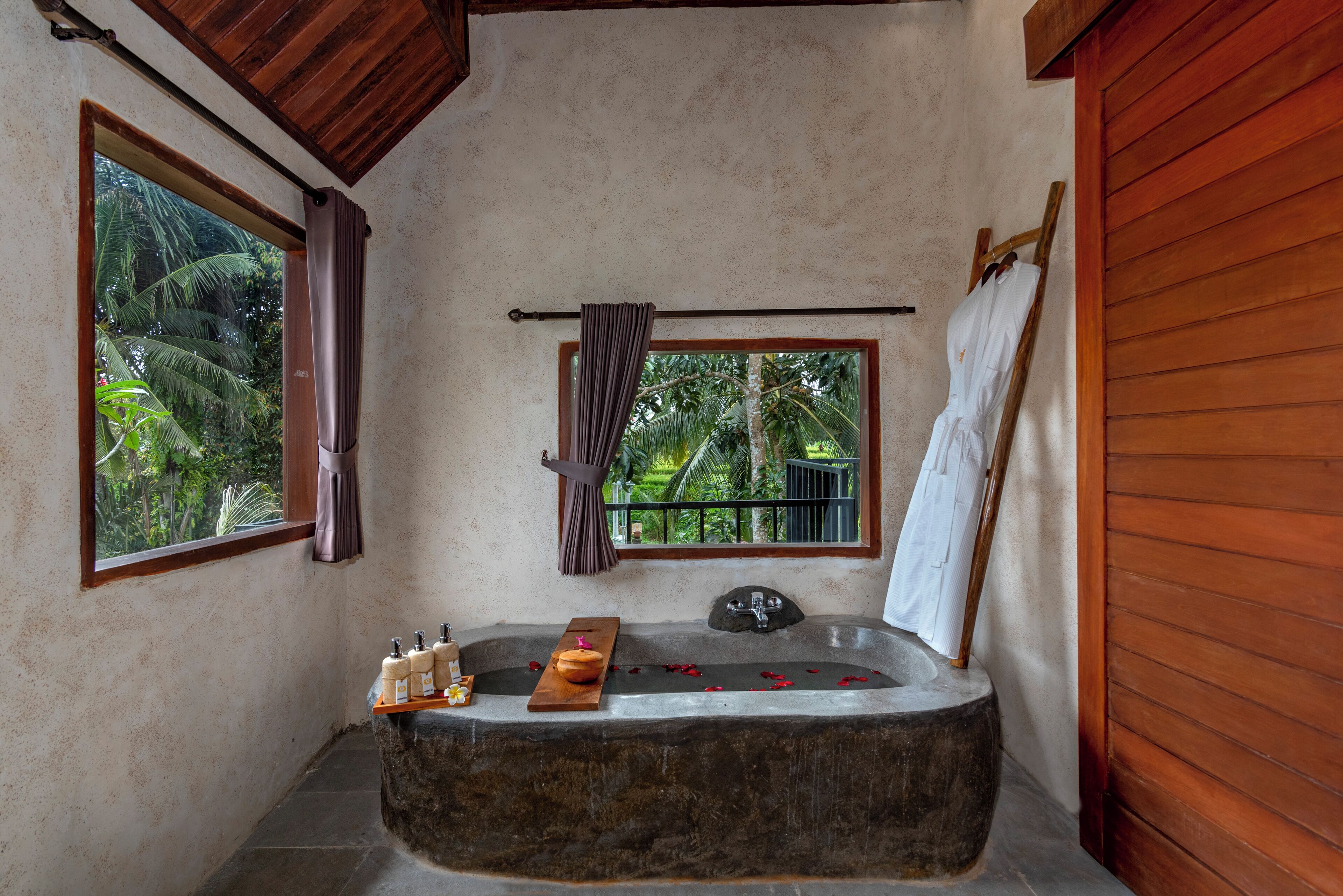 A bathroom with a stone bathtub, a wooden shelf with toiletries, a window with curtains, and a view of greenery outside.