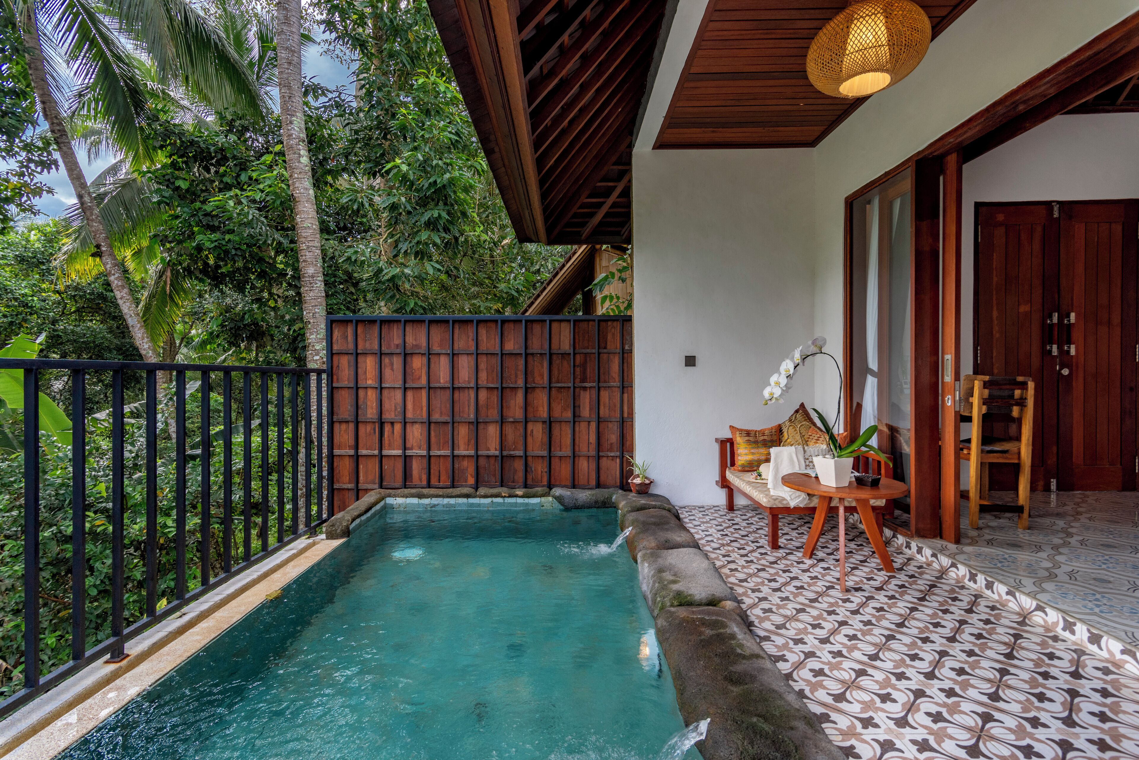 A pool area with a wooden deck, a seating area with a table and chairs, and a view of lush greenery.