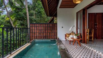 A pool area with a wooden deck, a seating area with a table and chairs, and a view of lush greenery.