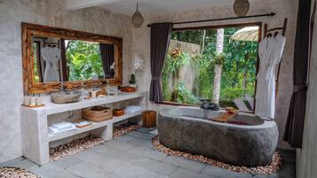 Casa de banho com uma banheira grande, toucador de madeira, espelho amplo e vista para o exterior, com vegetação.