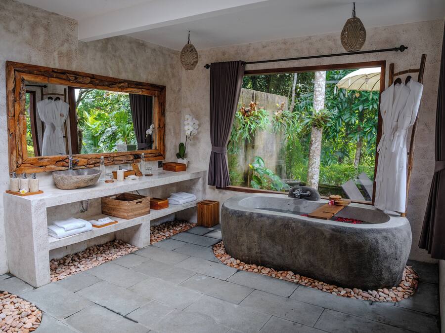 Casa de banho com uma banheira grande, toucador de madeira, espelho amplo e vista para o exterior, com vegetação.