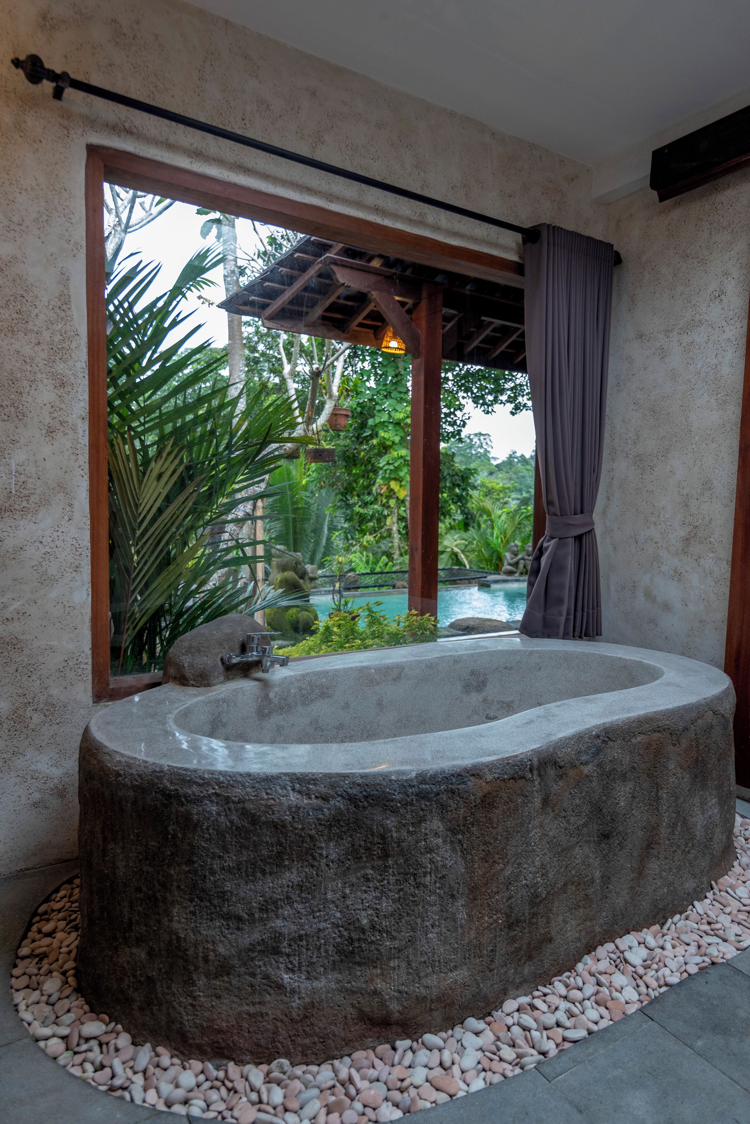 A large stone bathtub in a room with a view of a pool and greenery.