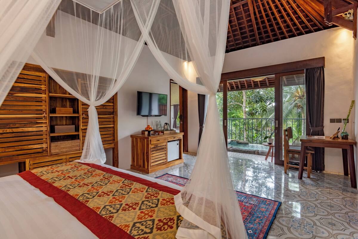 A bedroom with a bed, a wooden cabinet, a television, and a balcony with a view of greenery.