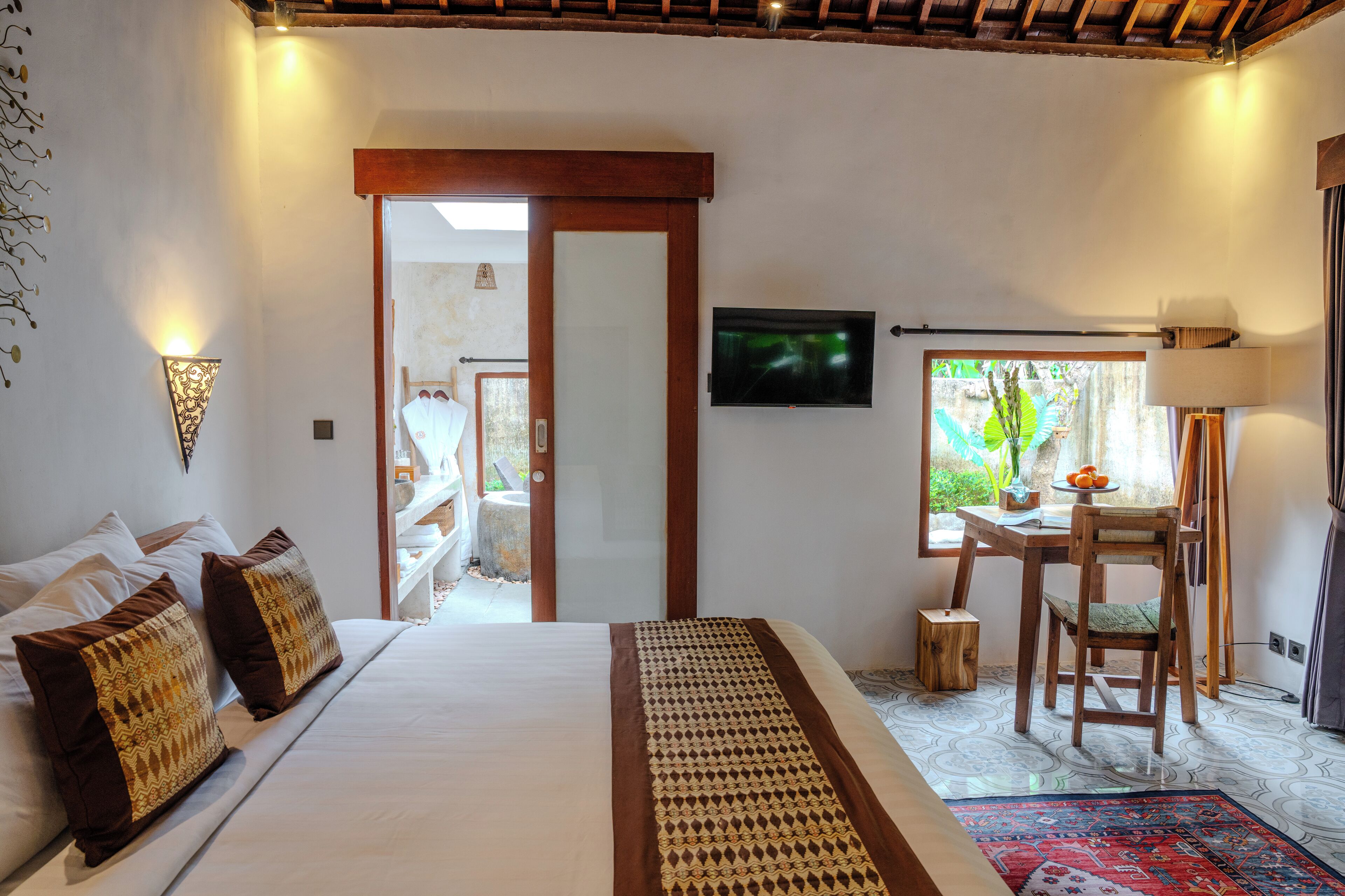 A bedroom with a bed, pillows, a TV, a desk with a chair, and a window with a view of greenery.