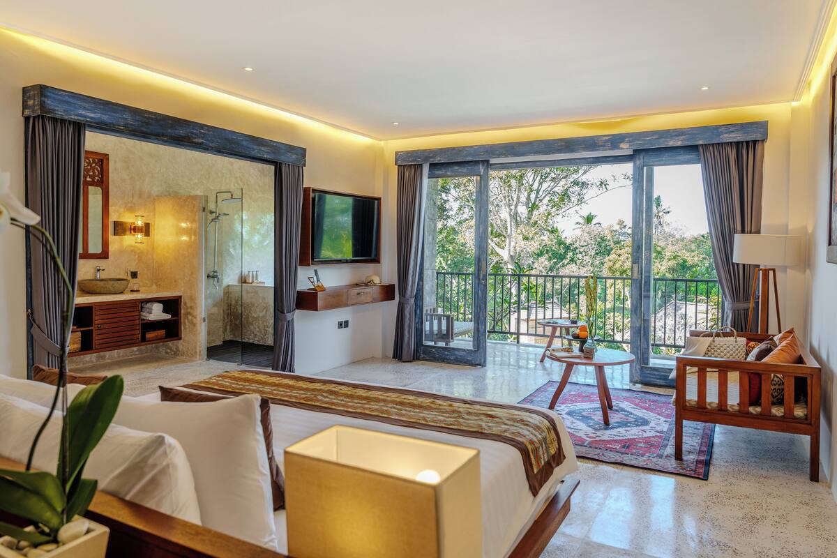 A modern hotel room with a large bed, a sitting area with a sofa and armchair, a small table, a TV, a bathroom with a shower, and a balcony with a view of trees.