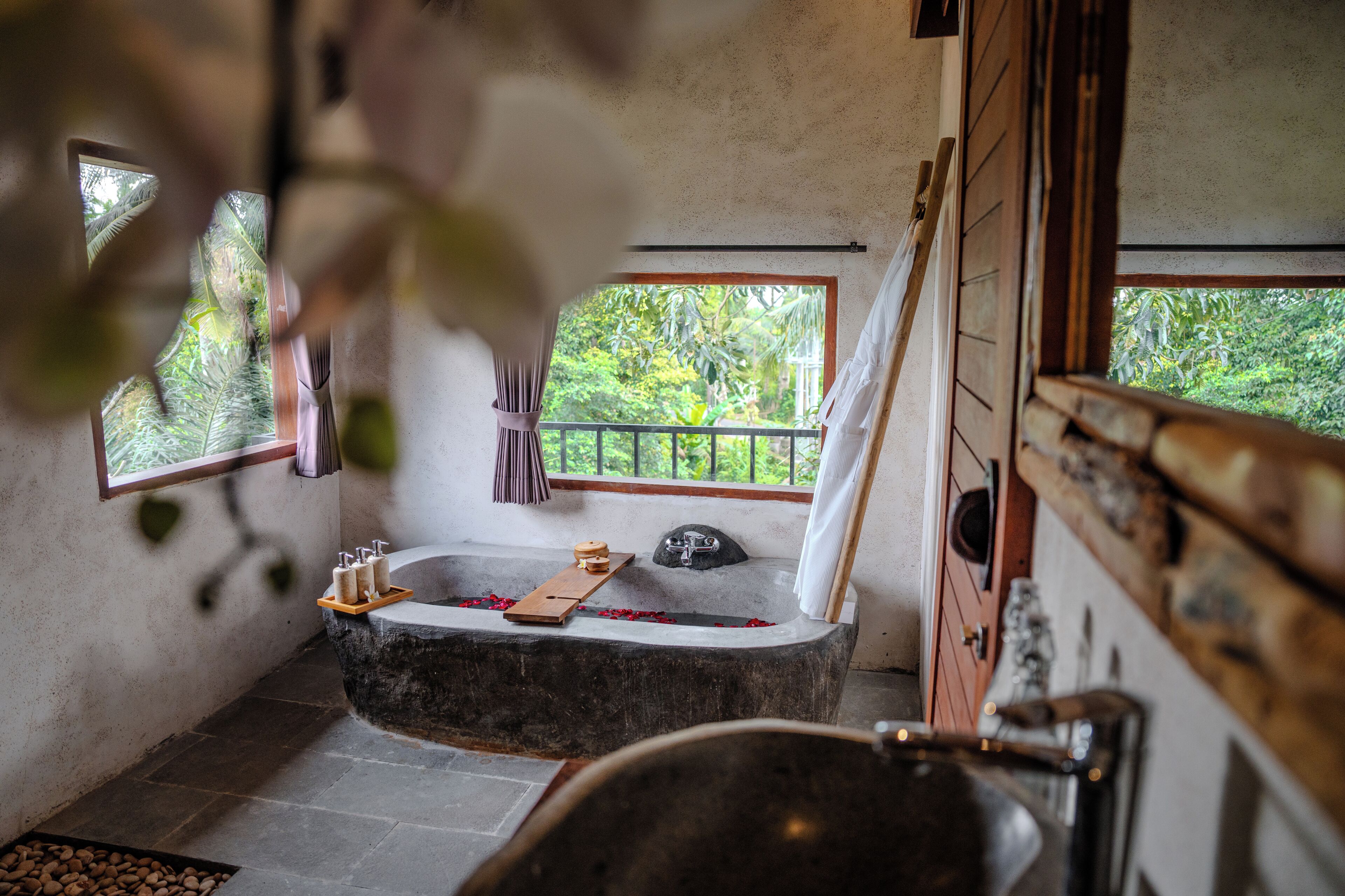 A bathroom with a stone bathtub, a wooden tray, and a window with a view of greenery.