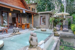 Un área de piscina con esculturas de piedra, una terraza de madera y una zona de estar cubierta con sombrillas.