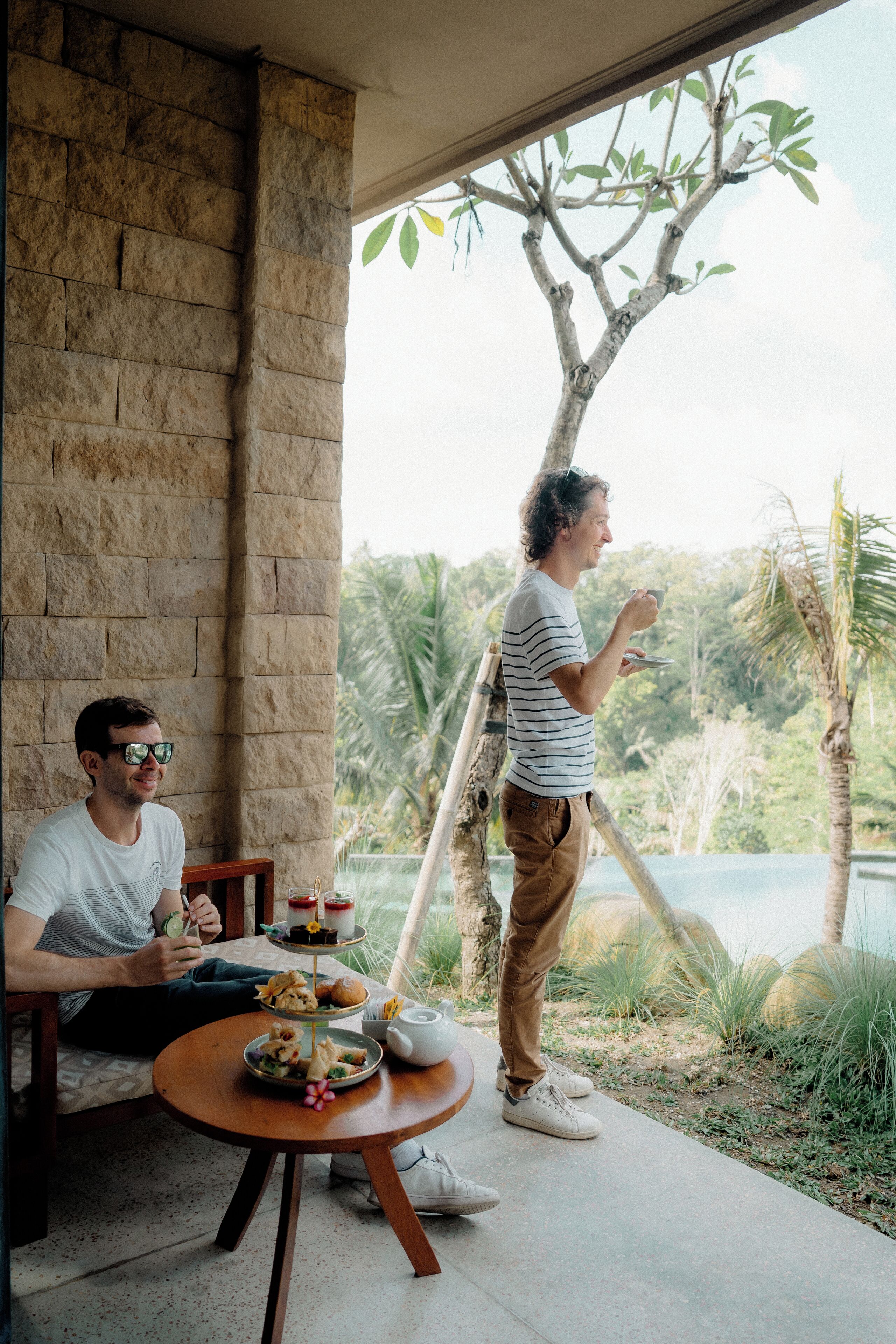 Un hombre de pie junto a una mesa con comida y una bebida, contemplando un espacio exterior con vistas a un cuerpo de agua y vegetación.