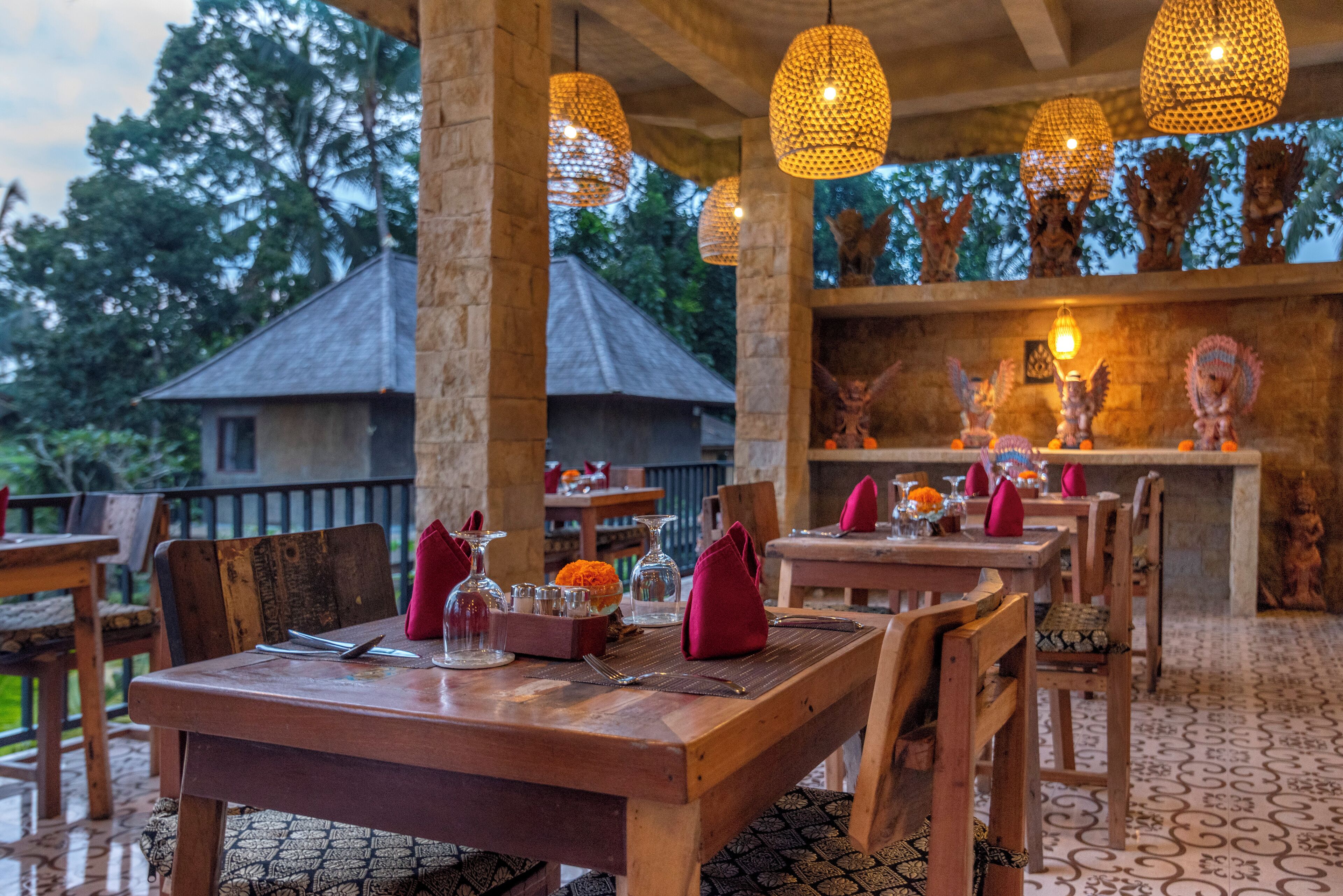 An outdoor dining area with wooden tables and chairs, hanging wicker lamps, and a stone wall with statues.