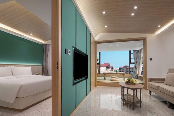 A hotel room with a bed, a TV, a sofa, a small table with fruit, and a view of buildings.
