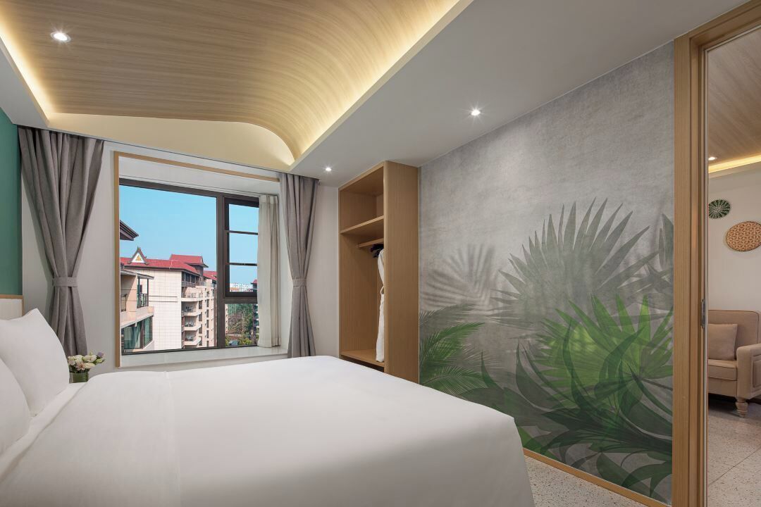 A hotel room with a large bed, a view of buildings through a window, a wooden ceiling, and a tropical mural on the wall.