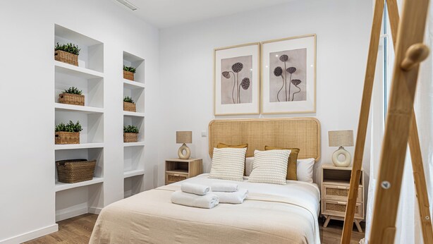 Une chambre avec un grand lit, deux tables de chevet, un plancher en bois, des murs blancs et des étagères avec des paniers et des plantes.