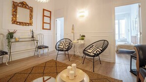 A well-lit living room with a large mirror, two black wire chairs, a small side table with candles, and a view into the bedroom.