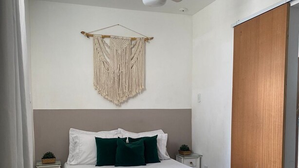 Un dormitorio con una cama, ventilador de techo, un tapiz de macramé, puerta de madera y una mesita de noche con una planta.