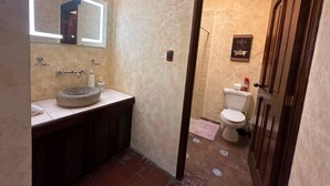 A bathroom with a stone sink, a mirror with lights, a toilet, and a door leading to another room. - Casa Los Héroes (Antigua Guatemala)