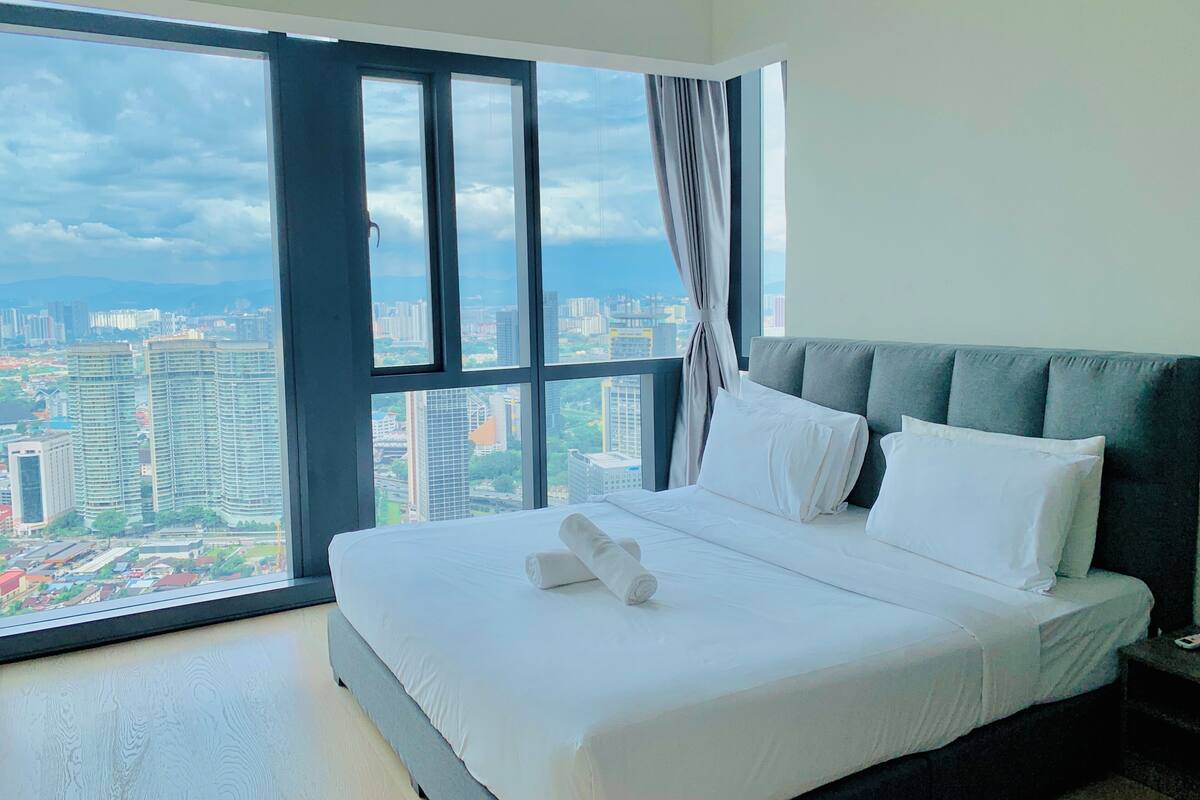 A hotel room with a large bed, a grey headboard, and a view of a cityscape through a large window.