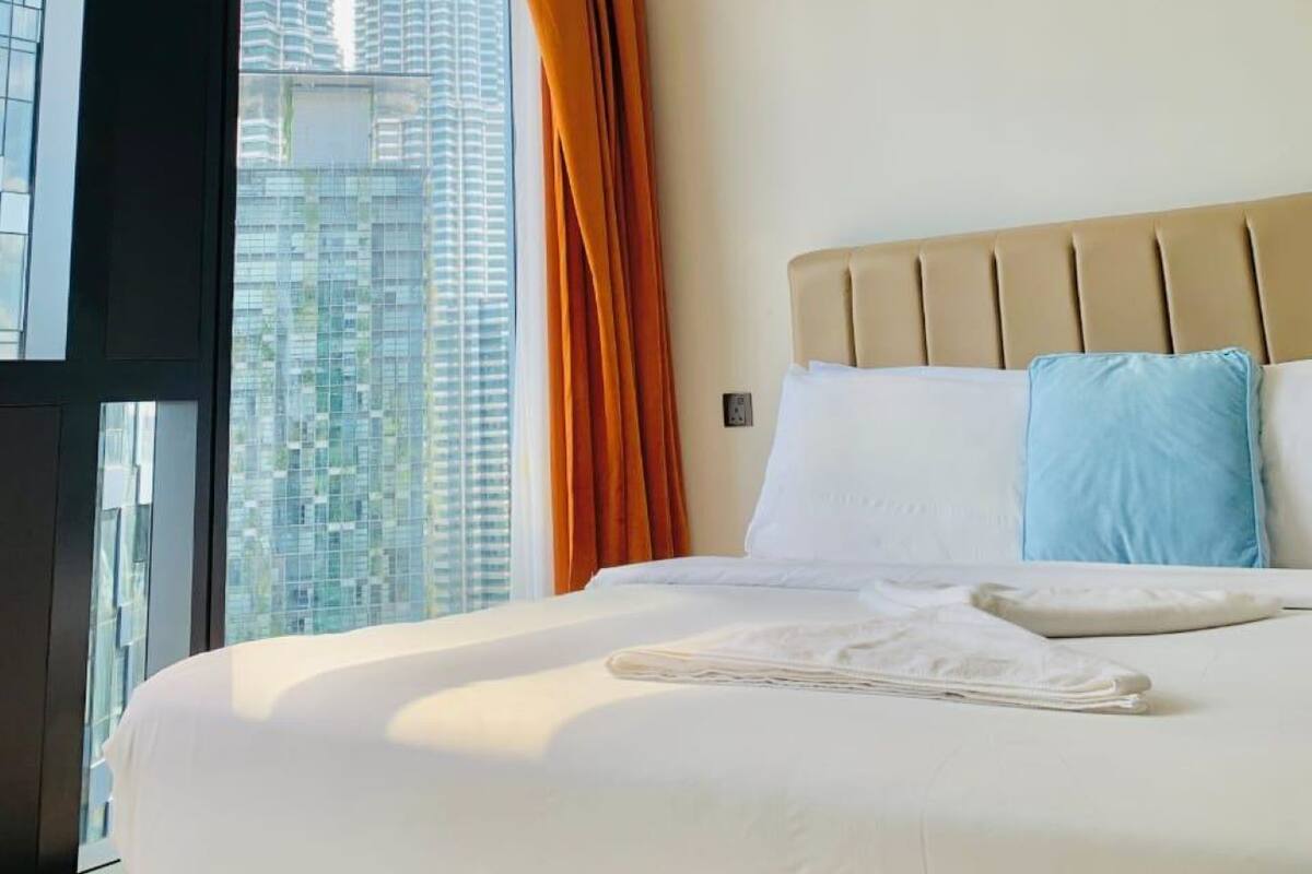 A hotel room with a large bed, a view of tall buildings, and a window with orange curtains.