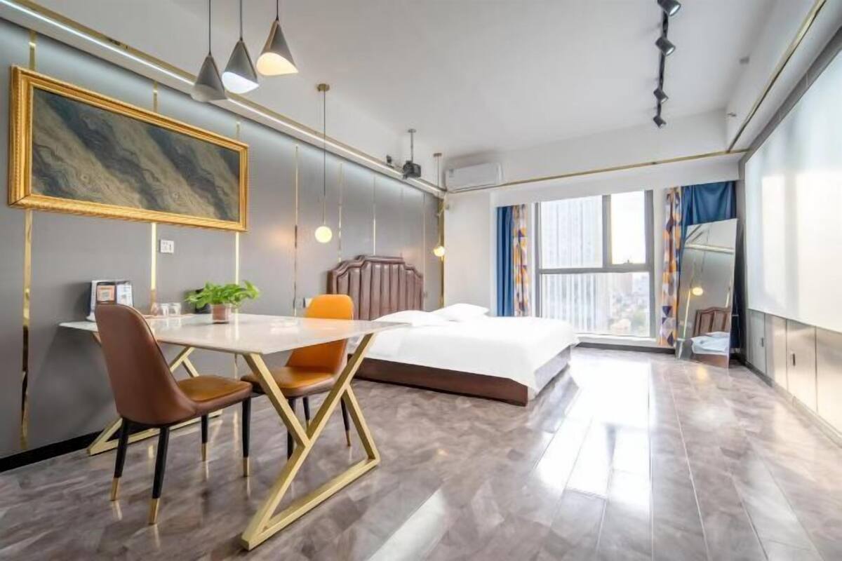 A modern hotel room with a large bed, a dining table with chairs, a desk with a chair, and a large window with curtains.