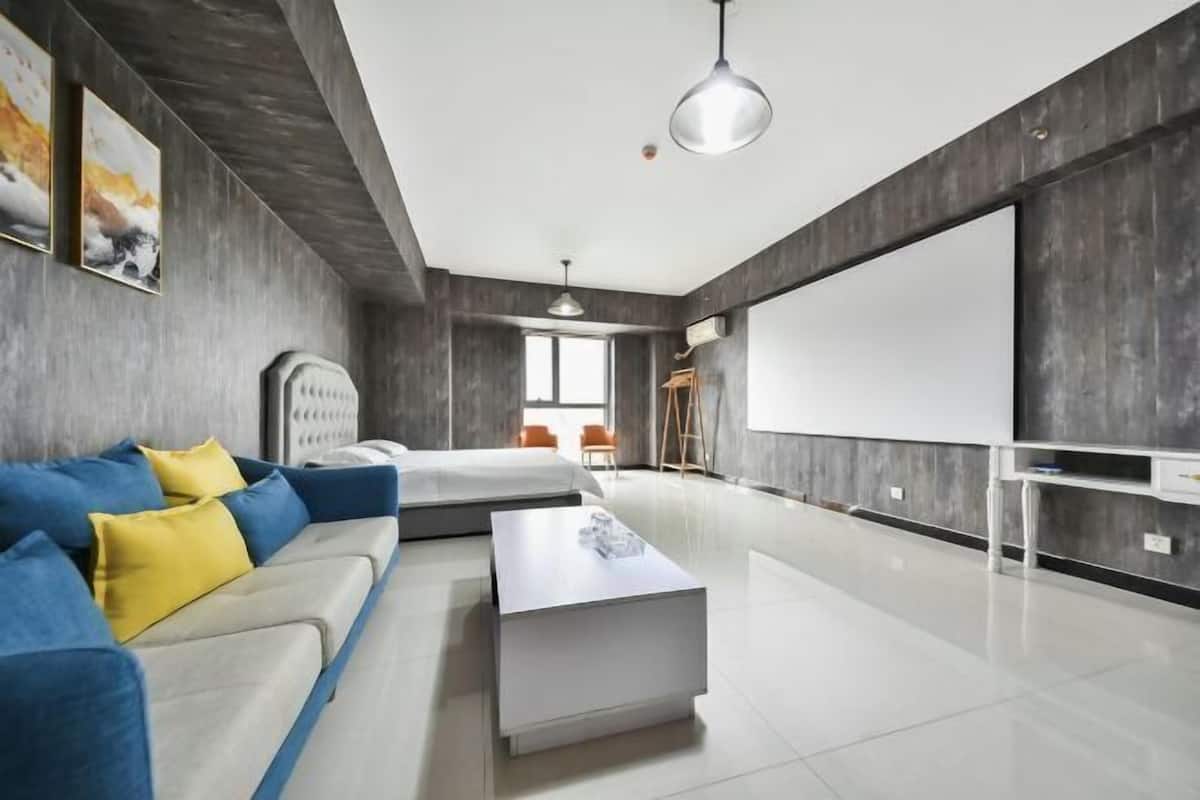 A modern hotel room with a large bed, a sofa with yellow cushions, a white coffee table, and a large flat-screen TV.