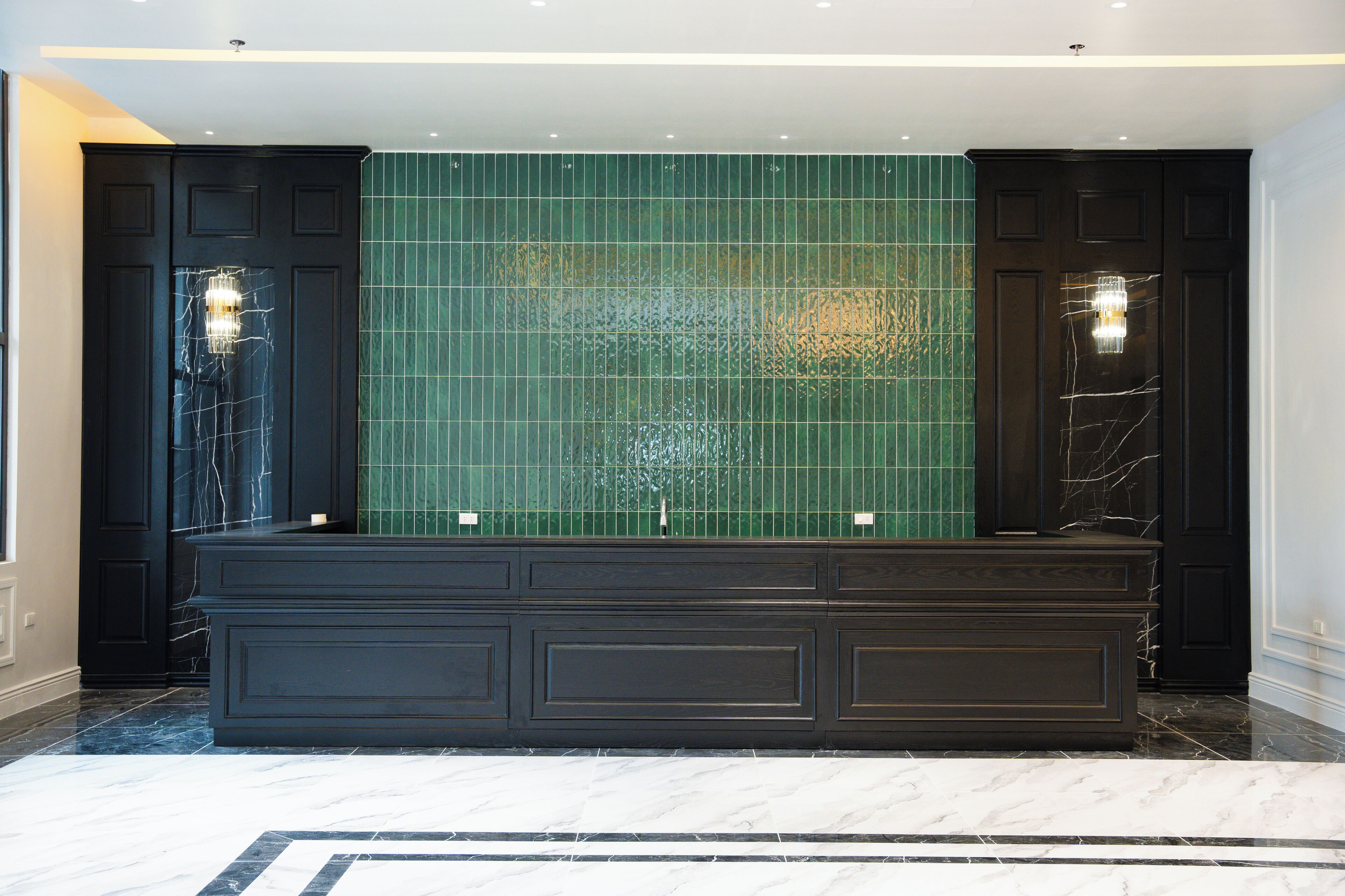 A dark-toned bathroom with a large mirror, green tiled wall, and two wall-mounted lights.