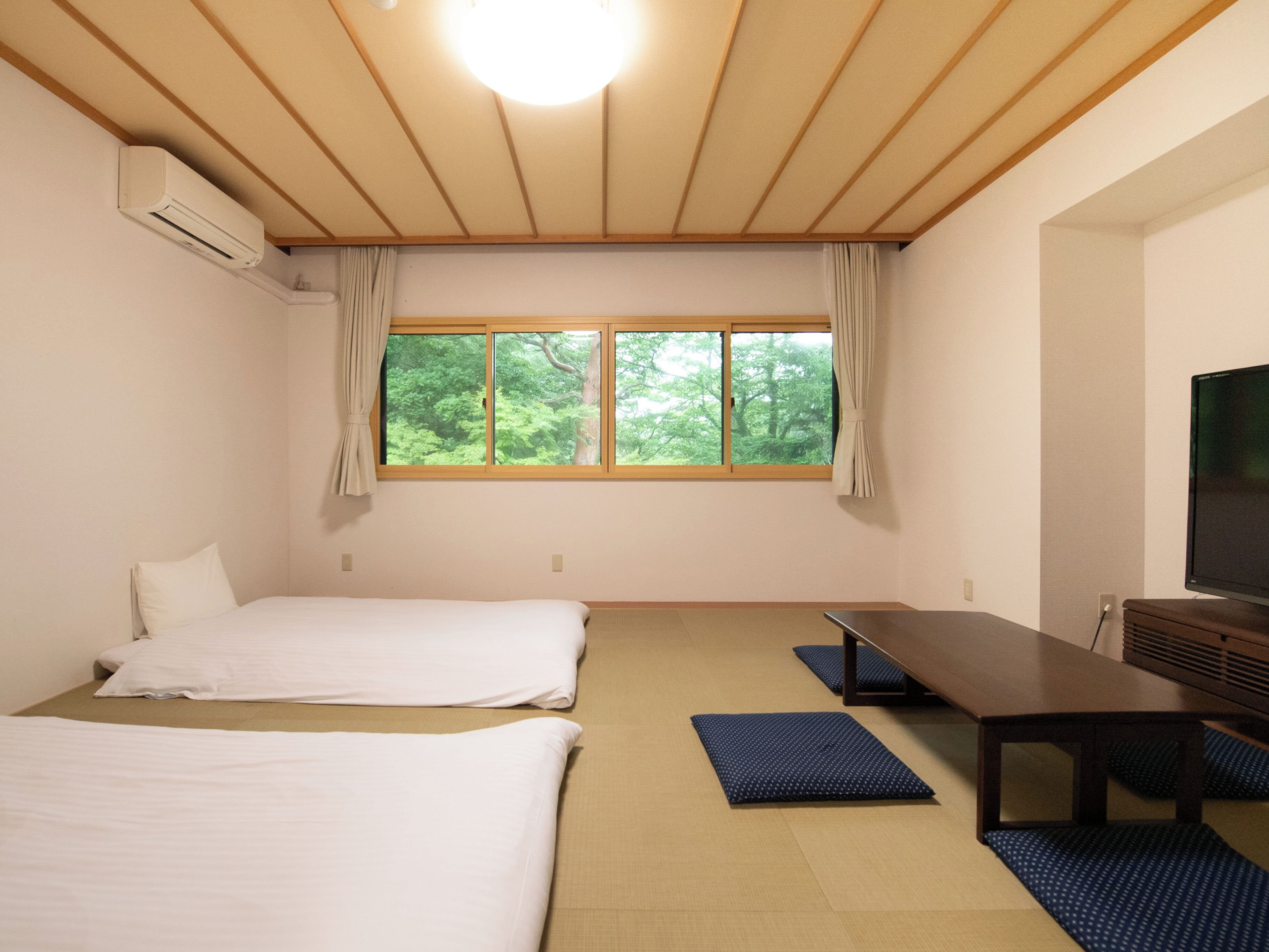 Een hotelkamer met twee bedden, een televisie, een raam met uitzicht op bomen, en een plafondventilator.
