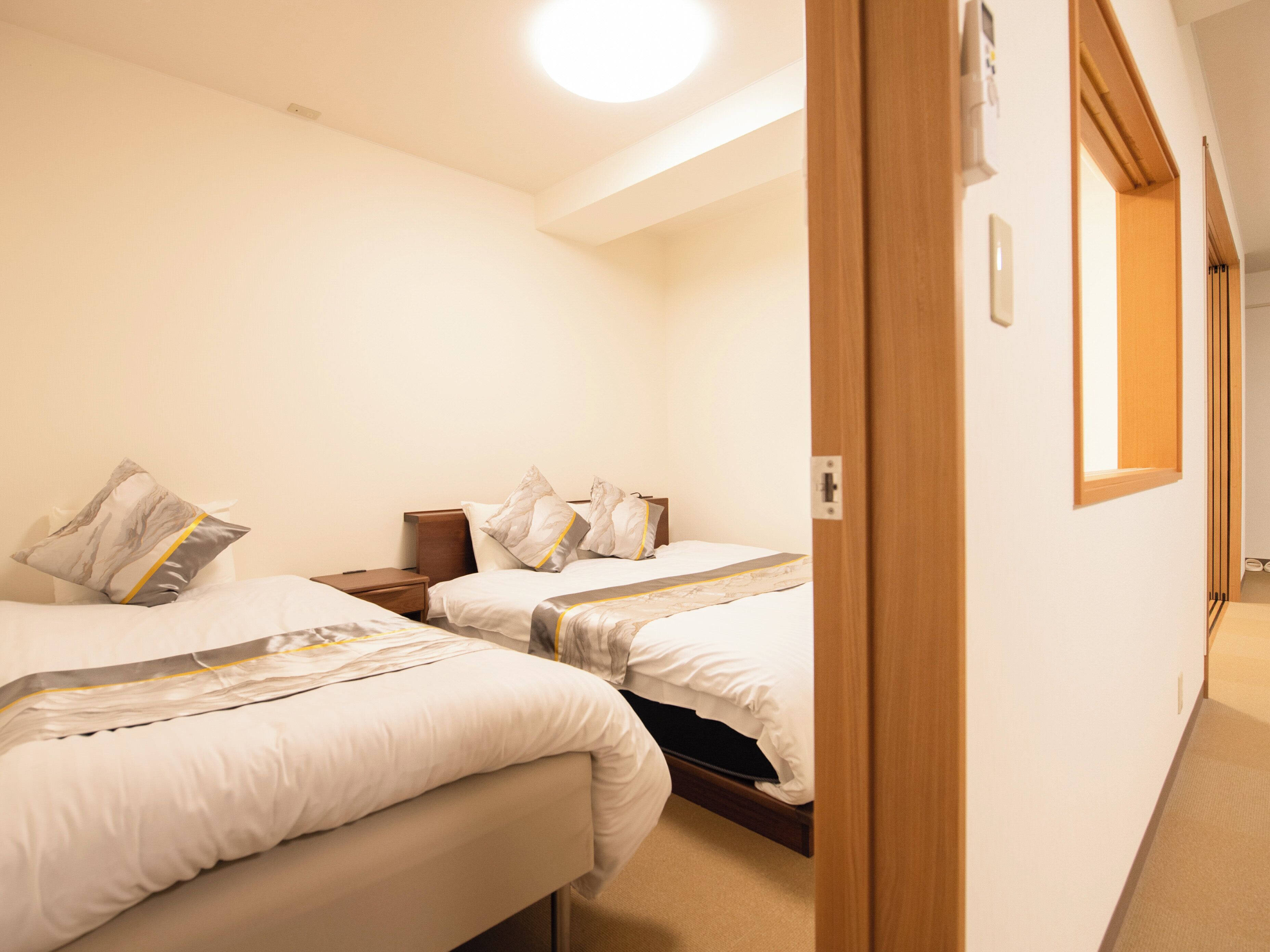 Une chambre d’hôtel avec deux lits, une porte en bois, une petite table de chevet et un luminaire au plafond.