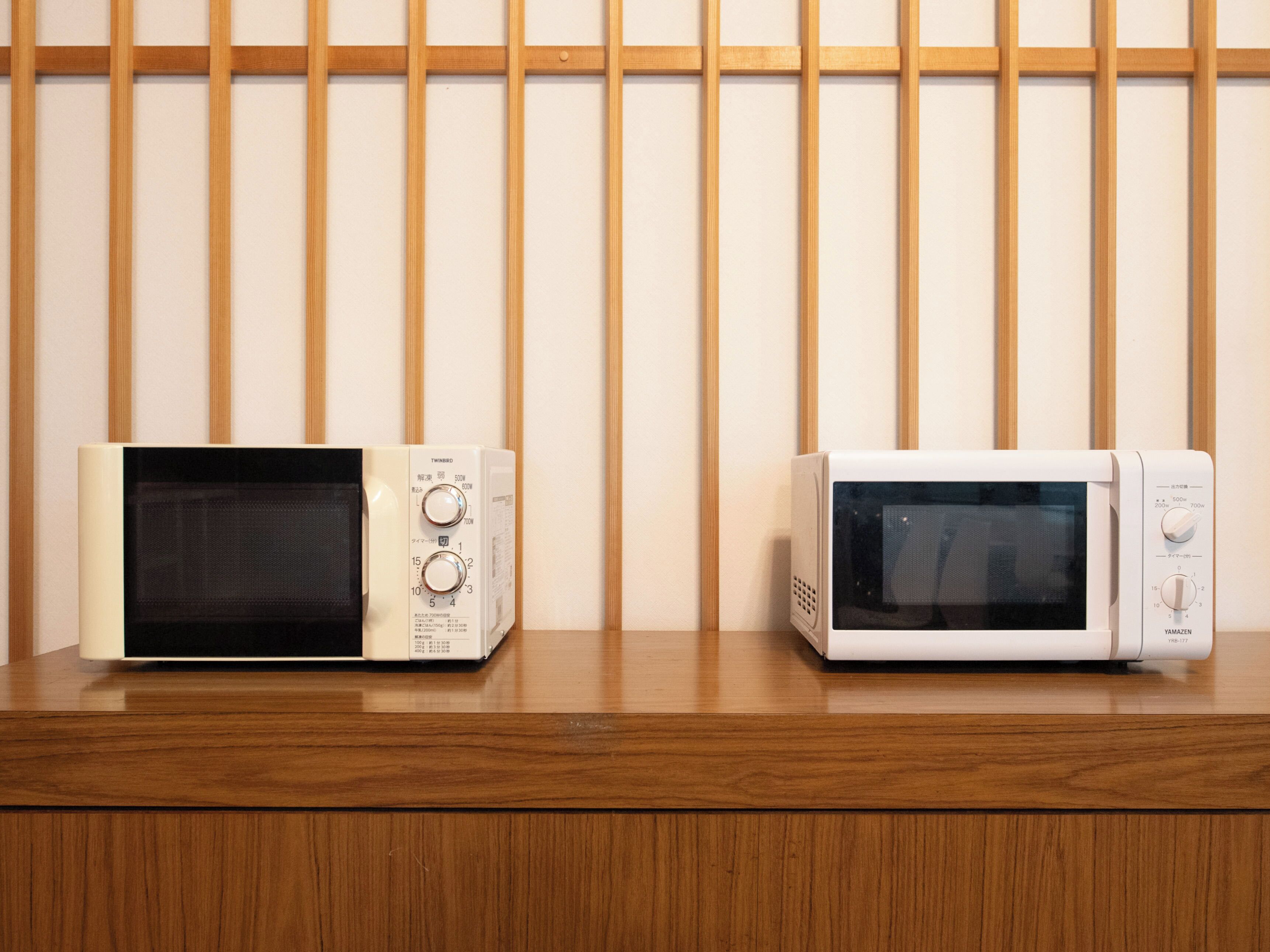 Two microwaves on a wooden surface with a wooden slat wall in the background.