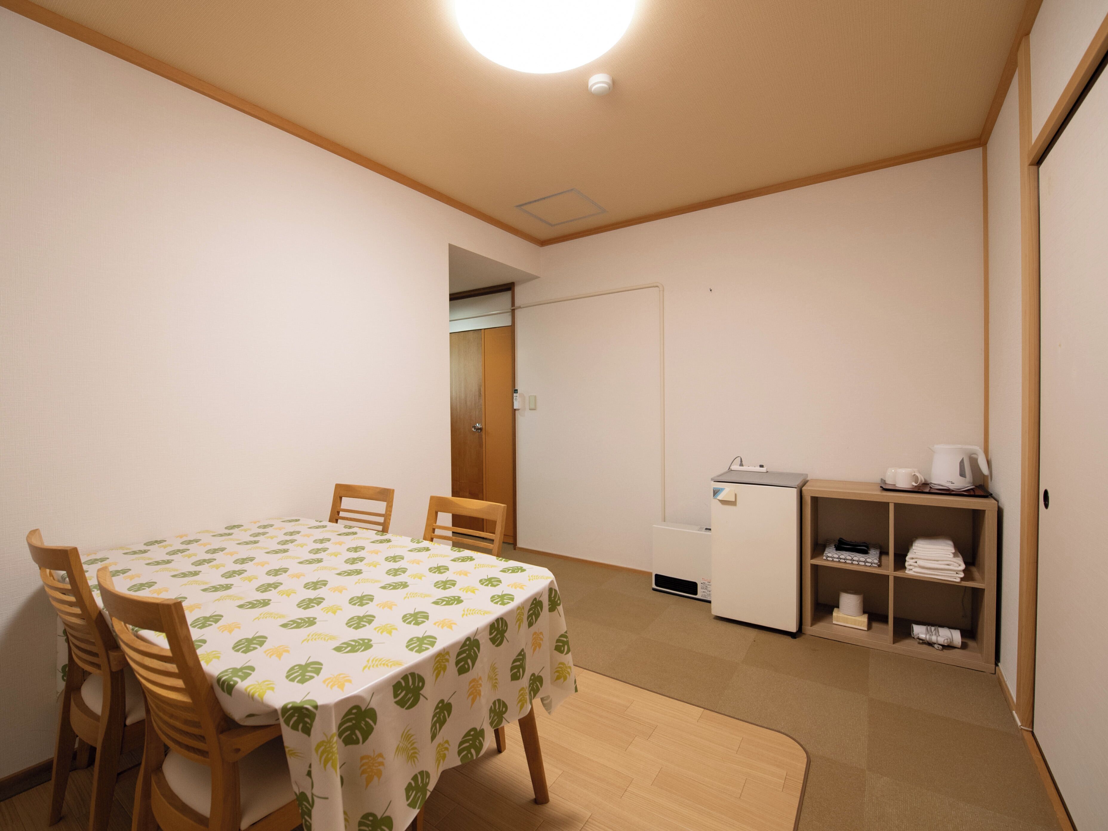 A room with a table covered in a leaf-patterned tablecloth, wooden chairs, a refrigerator, a shelf with towels, and a door leading to another room.
